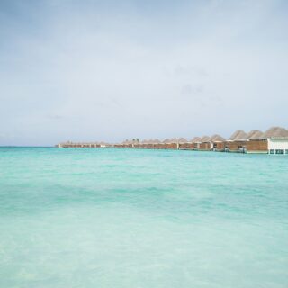 Overwater bungalows with thatched roofs line a turquoise lagoon under a clear sky.