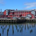 Red and gray waterfront hotel built on stilts over calm blue water, with a wooden boardwalk in front and a clear sky above.