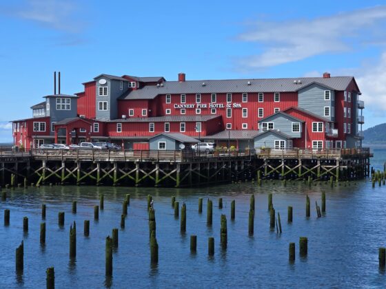 Red and gray waterfront hotel built on stilts over calm blue water, with a wooden boardwalk in front and a clear sky above.