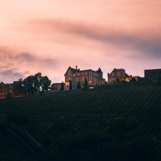 Hilltop hotel complex with castle-like buildings at sunset, purple-pink sky and terraced slope below.