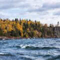 Lighthouse perched on a rocky cliff with colorful autumn trees along the shore and choppy water in the foreground.
