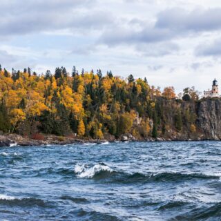 Lighthouse perched on a rocky cliff with colorful autumn trees along the shore and choppy water in the foreground.