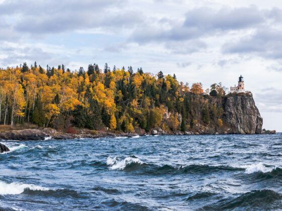 Lighthouse perched on a rocky cliff with colorful autumn trees along the shore and choppy water in the foreground.