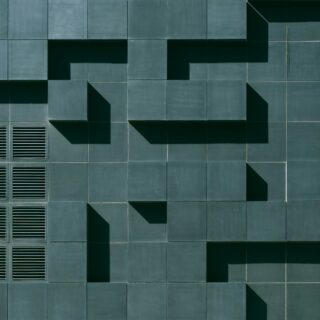 Teal tiled building facade with square panels and deep geometric shadows forming stair-step patterns.