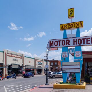 Yellow Route 66 sign reading ARIZONA with a small '9' on top, supported by blue posts under a clear blue sky, near a motor hotel sign.