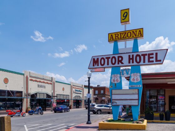 Yellow Route 66 sign reading ARIZONA with a small '9' on top, supported by blue posts under a clear blue sky, near a motor hotel sign.