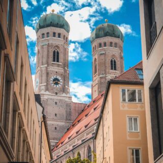 Two church towers with green domes rise over a narrow street lined with beige buildings against a blue sky with clouds.