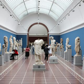 Long museum gallery with white statues on pedestals along a central aisle, blue walls, and a glass arched ceiling.