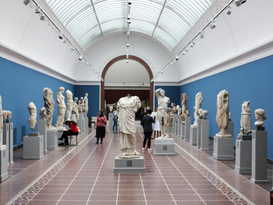 Long museum gallery with white statues on pedestals along a central aisle, blue walls, and a glass arched ceiling.