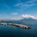 Harbor scene with a breakwater of rocks, calm blue water, and distant mountains under a clear sky with scattered clouds