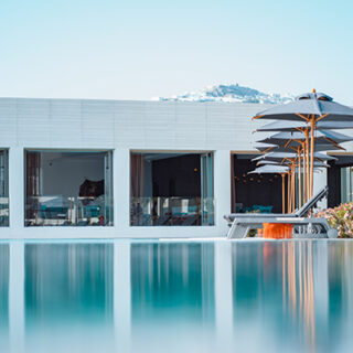 Poolside at a modern white resort: glass-walled rooms, lounge chairs, and stacked umbrellas along a calm reflective pool.