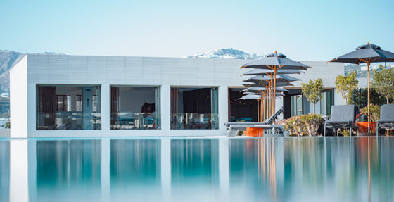 Poolside at a modern white resort: glass-walled rooms, lounge chairs, and stacked umbrellas along a calm reflective pool.