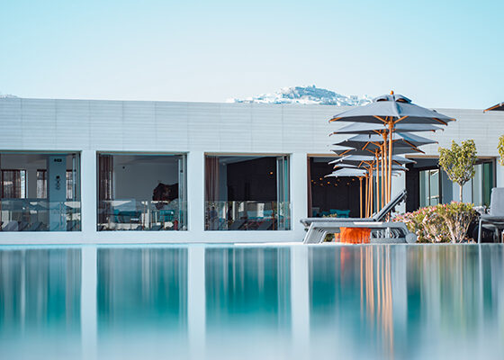 Poolside at a modern white resort: glass-walled rooms, lounge chairs, and stacked umbrellas along a calm reflective pool.