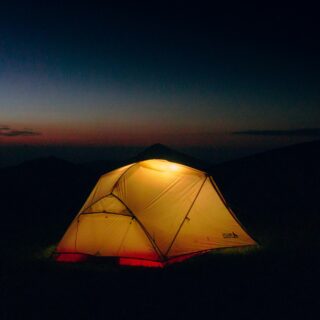 Lit yellow camping tent glowing at night, set on a dark hillside with a dusky gradient sky and distant mountains in the background.