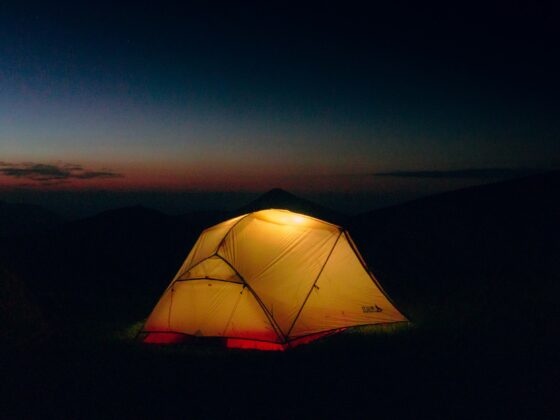 Lit yellow camping tent glowing at night, set on a dark hillside with a dusky gradient sky and distant mountains in the background.