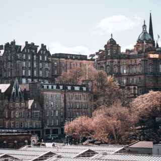 Panoramic view of Edinburgh’s historic brick buildings, a domed tower and tall spire rise above, with a glass-roofed foreground and pink trees.