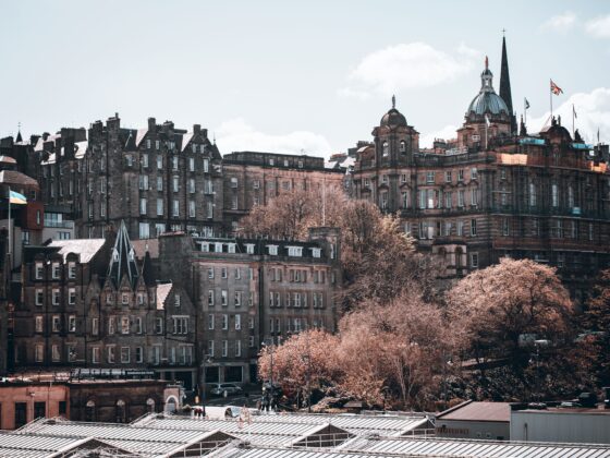 Panoramic view of Edinburgh’s historic brick buildings, a domed tower and tall spire rise above, with a glass-roofed foreground and pink trees.