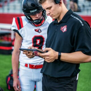 Two football players on the sideline, one in a black polo with a headset looking at a tablet while the other wears a white uniform and helmet nearby on the field edge.