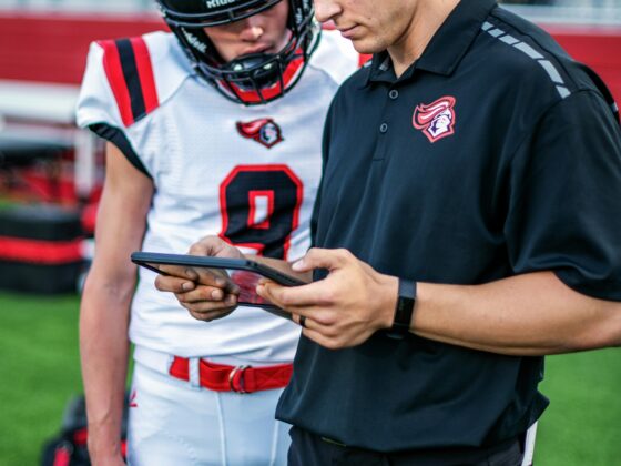 Two football players on the sideline, one in a black polo with a headset looking at a tablet while the other wears a white uniform and helmet nearby on the field edge.