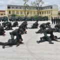 Army trainees in green uniforms perform a ground drill: crawling and grappling on a wide parade ground, with trucks and a building in the background.