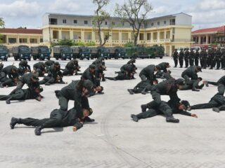 Army trainees in green uniforms perform a ground drill: crawling and grappling on a wide parade ground, with trucks and a building in the background.