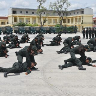 Army trainees in green uniforms perform a ground drill: crawling and grappling on a wide parade ground, with trucks and a building in the background.