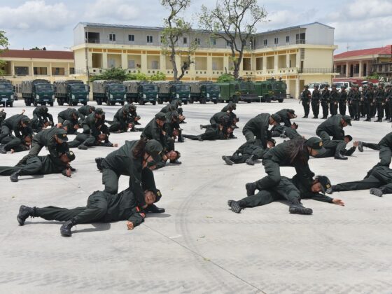 Army trainees in green uniforms perform a ground drill: crawling and grappling on a wide parade ground, with trucks and a building in the background.