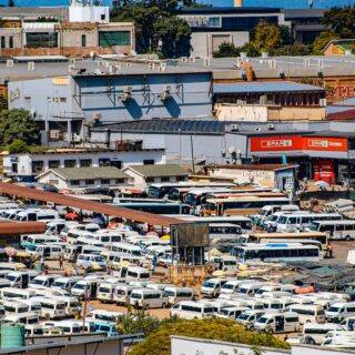 Large outdoor parking lot filled with dozens of white vans next to market stalls and industrial buildings under a blue sky.