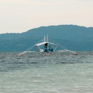 Traditional boat with white masts floating on calm blue-green water, hills in the distance under a pale sky.