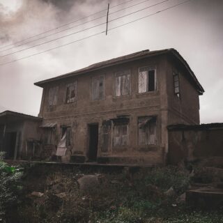 Dilapidated two-story house with broken windows and weathered walls, overgrown yard and overhead power lines against a cloudy sky.