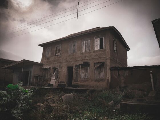 Dilapidated two-story house with broken windows and weathered walls, overgrown yard and overhead power lines against a cloudy sky.