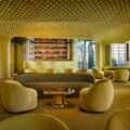 Modern lounge with pale yellow seating and a dotted ceiling, circular chairs around a shiny coffee table, bar area in back.