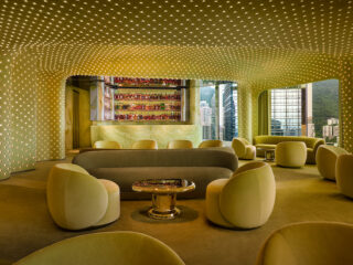 Modern lounge with pale yellow seating and a dotted ceiling, circular chairs around a shiny coffee table, bar area in back.
