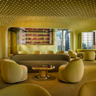 Modern lounge with pale yellow seating and a dotted ceiling, circular chairs around a shiny coffee table, bar area in back.