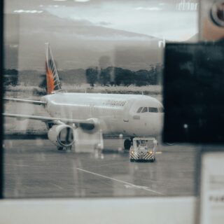Air passenger jet at the gate seen through a glass window, with the orange-tailed engine area visible on the tarmac.