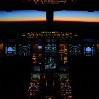 Dark airplane cockpit at night with orange-lit instruments and displays, sunset visible through the windshield ahead.