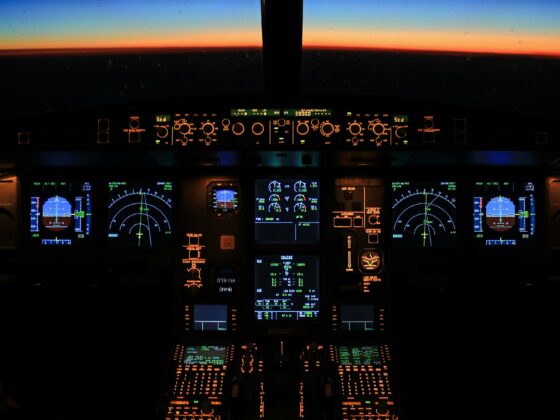 Dark airplane cockpit at night with orange-lit instruments and displays, sunset visible through the windshield ahead.
