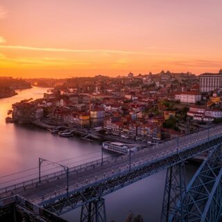 Sunset over a riverfront city with a large metal bridge crossing the water and colorful buildings along the shore.
