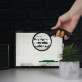 Close-up of a hand with orange nail polish gripping a device over a tablet displaying a circular sticker and partial text, against a dark brick wall background.