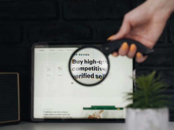 Close-up of a hand with orange nail polish gripping a device over a tablet displaying a circular sticker and partial text, against a dark brick wall background.