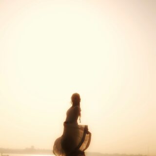 Silhouetted woman dancing on the beach at sunset, a flowing dress catching the breeze with a large sun behind.