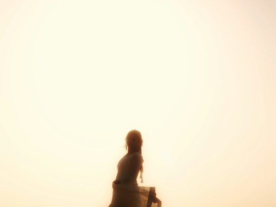 Silhouetted woman dancing on the beach at sunset, a flowing dress catching the breeze with a large sun behind.