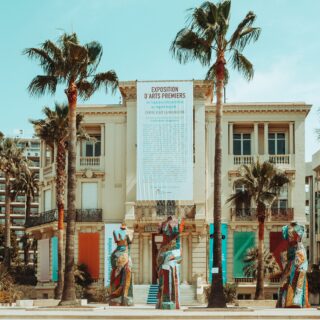 Beige neoclassical building with a tall banner displaying art, flanked by palm trees on a sunny day.