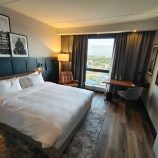 Hotel room with two white beds, dark wood headboard, and framed black-and-white art above the headboard; soft lighting from a floor lamp. A leather chair sits by a desk near a large window with city view and gray curtains.