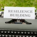 Vintage typewriter on a stone surface with a sheet reading RESILIENCE BUILDING, outdoors on grass.