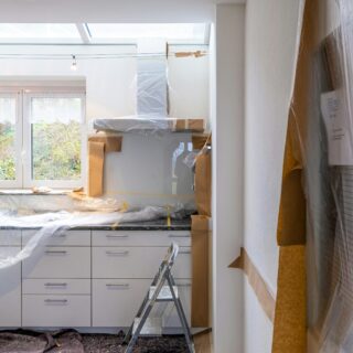 Kitchen under renovation with plastic sheeting over cabinets and appliances, and a metal step ladder in the center.