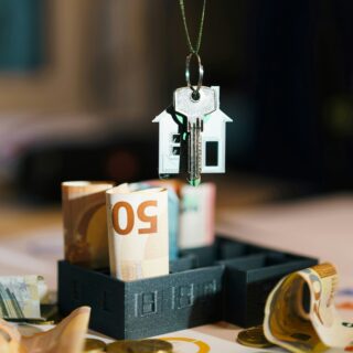 House key on a pendant shaped like a small house, hanging above rolled euro banknotes and scattered coins on a desk or table.