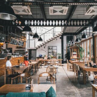 Bright cafe interior with wooden tables and chairs, black pendant lights, and large windows on the right; customers sit and work or chat.