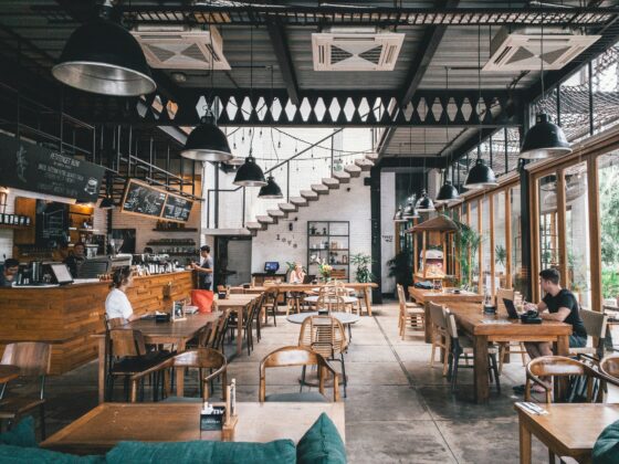 Bright cafe interior with wooden tables and chairs, black pendant lights, and large windows on the right; customers sit and work or chat.