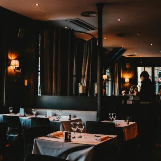 Dimly lit restaurant dining area with white-tablecloth tables, wine glasses, and candles along a dark, elegant room.